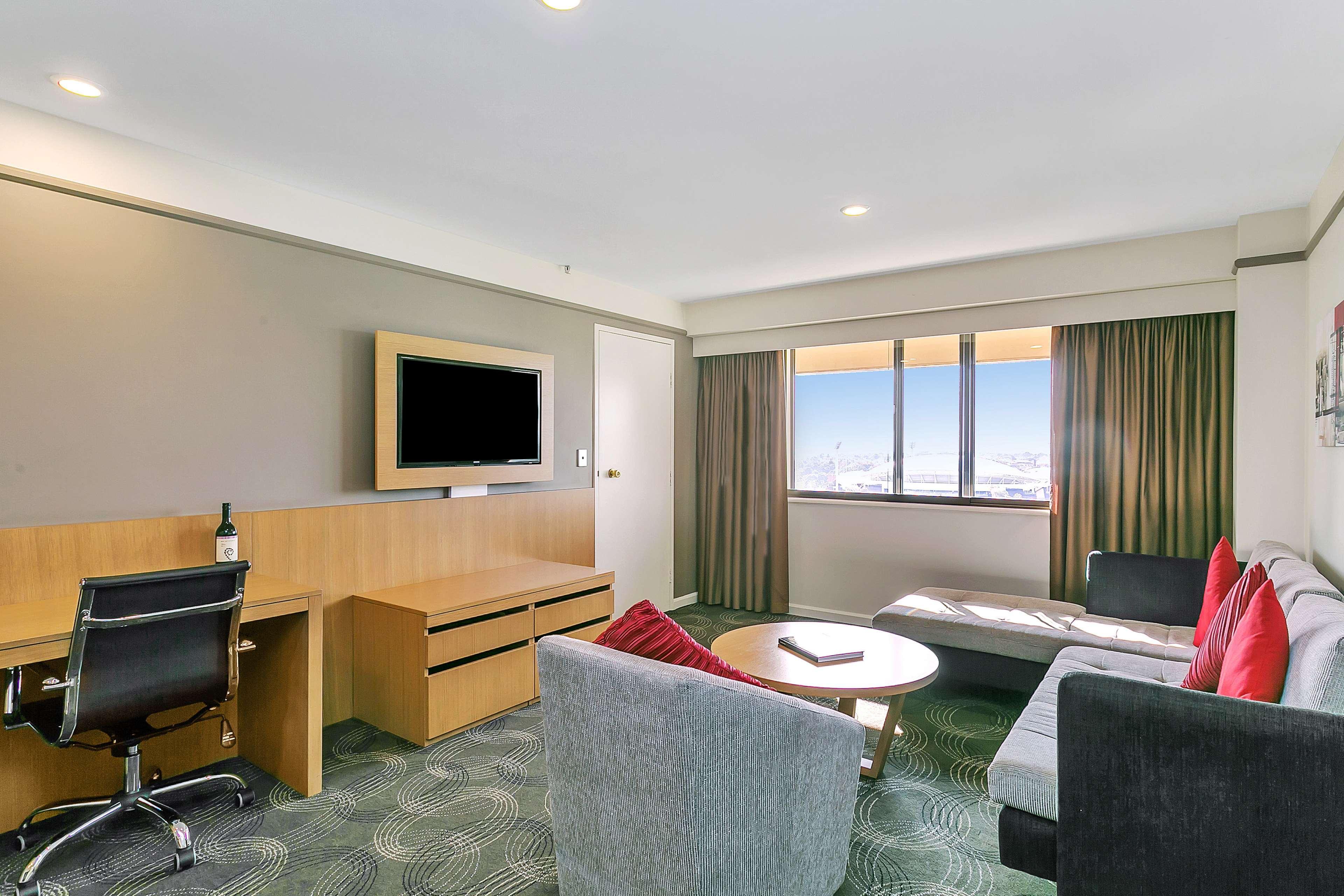 스탬퍼드 플라자 애들레이드 호텔 외부 사진 The photo shows a modern hotel room or suite that features a cozy seating area. There is a comfortable sofa with red accent pillows, a round coffee table in the center, and a sleek desk with an office chair. On the wall, there is a flat-screen TV mou