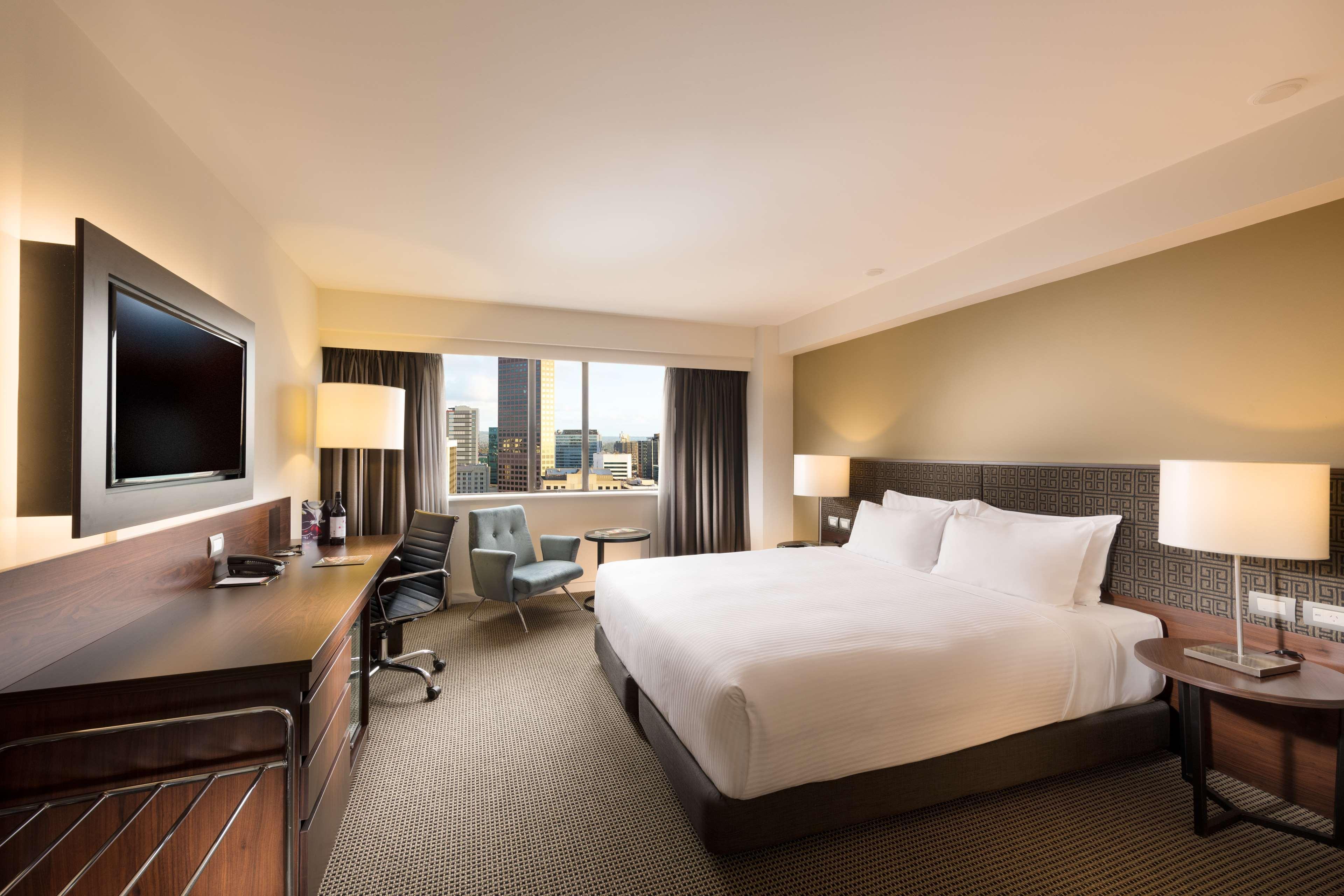 스탬퍼드 플라자 애들레이드 호텔 외부 사진 The photo shows a modern hotel room with a contemporary design. It features a large bed with white linens, a bedside table on either side with lamps, and a dark wood accent wall. There's a work desk with an office chair and a flat-screen television m