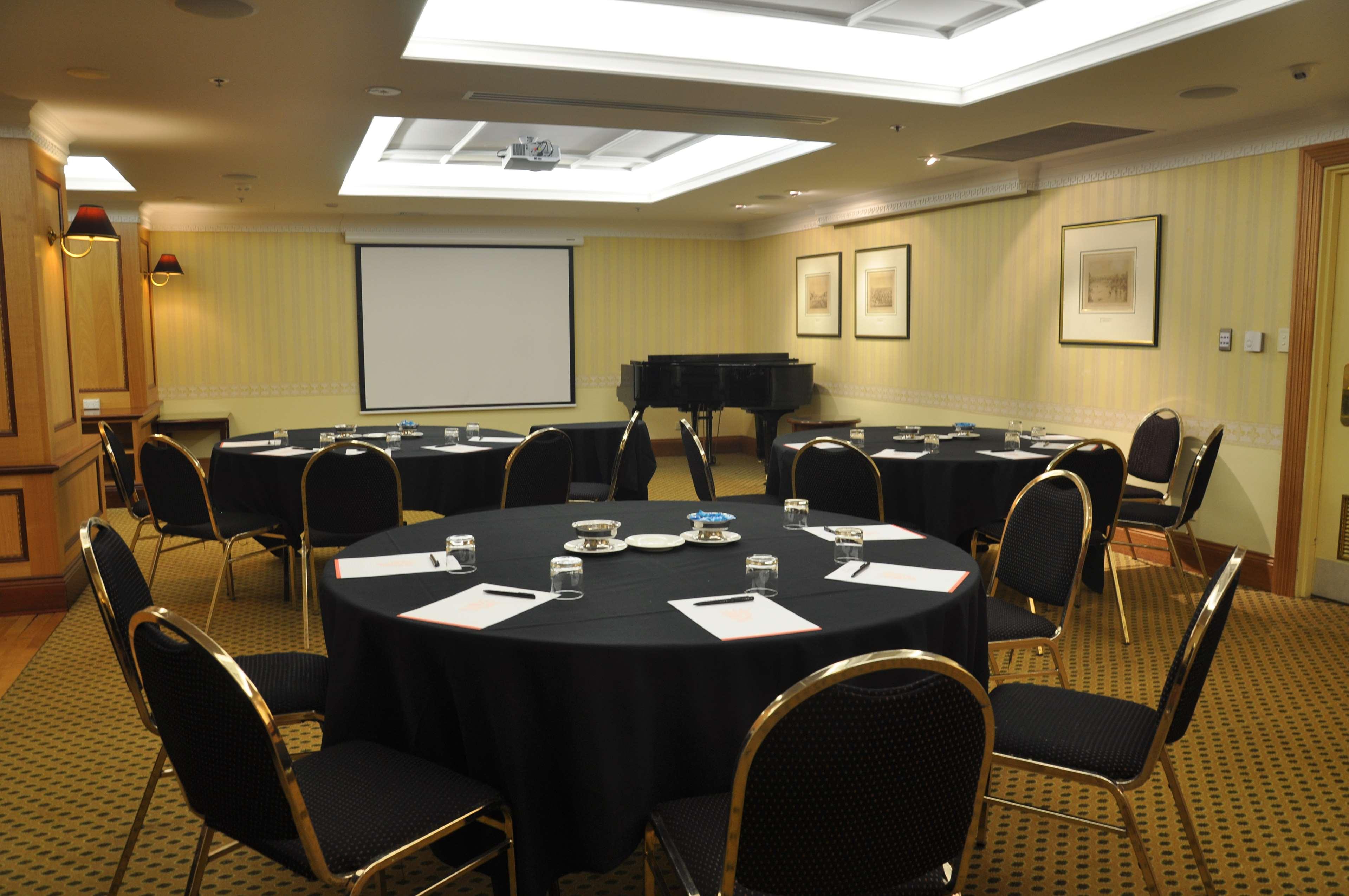 스탬퍼드 플라자 애들레이드 호텔 외부 사진 The photo shows a conference room set up for a meeting or presentation. There are several round tables covered with black tablecloths, each surrounded by black upholstered chairs. On the tables, there are notepads and glasses of water, indicating tha