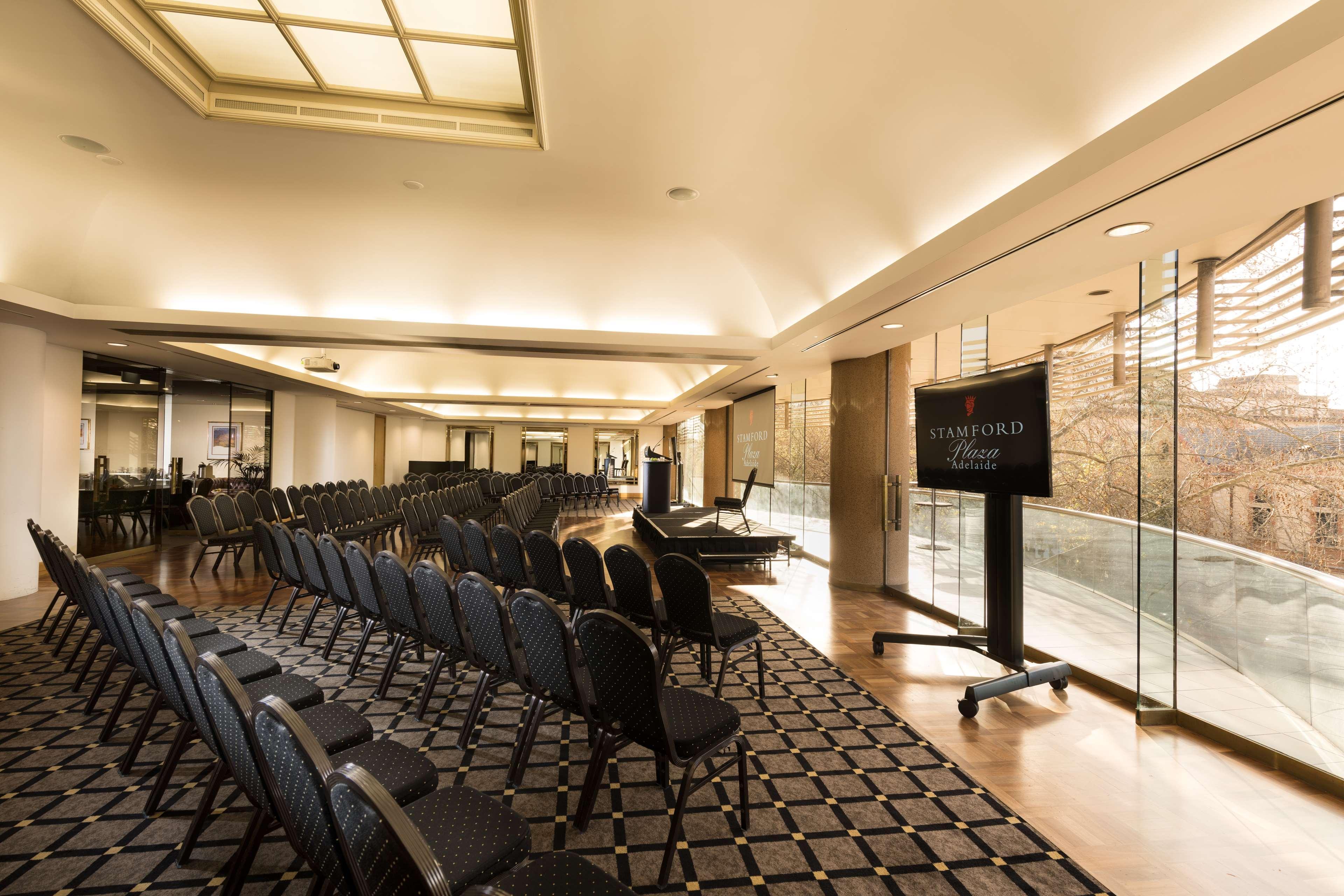 스탬퍼드 플라자 애들레이드 호텔 외부 사진 The photo shows a large, elegantly designed event space or meeting room. There are rows of black chairs set up facing a presentation area. The flooring features a patterned carpet, and large windows allow natural light to enter the room. On one side,