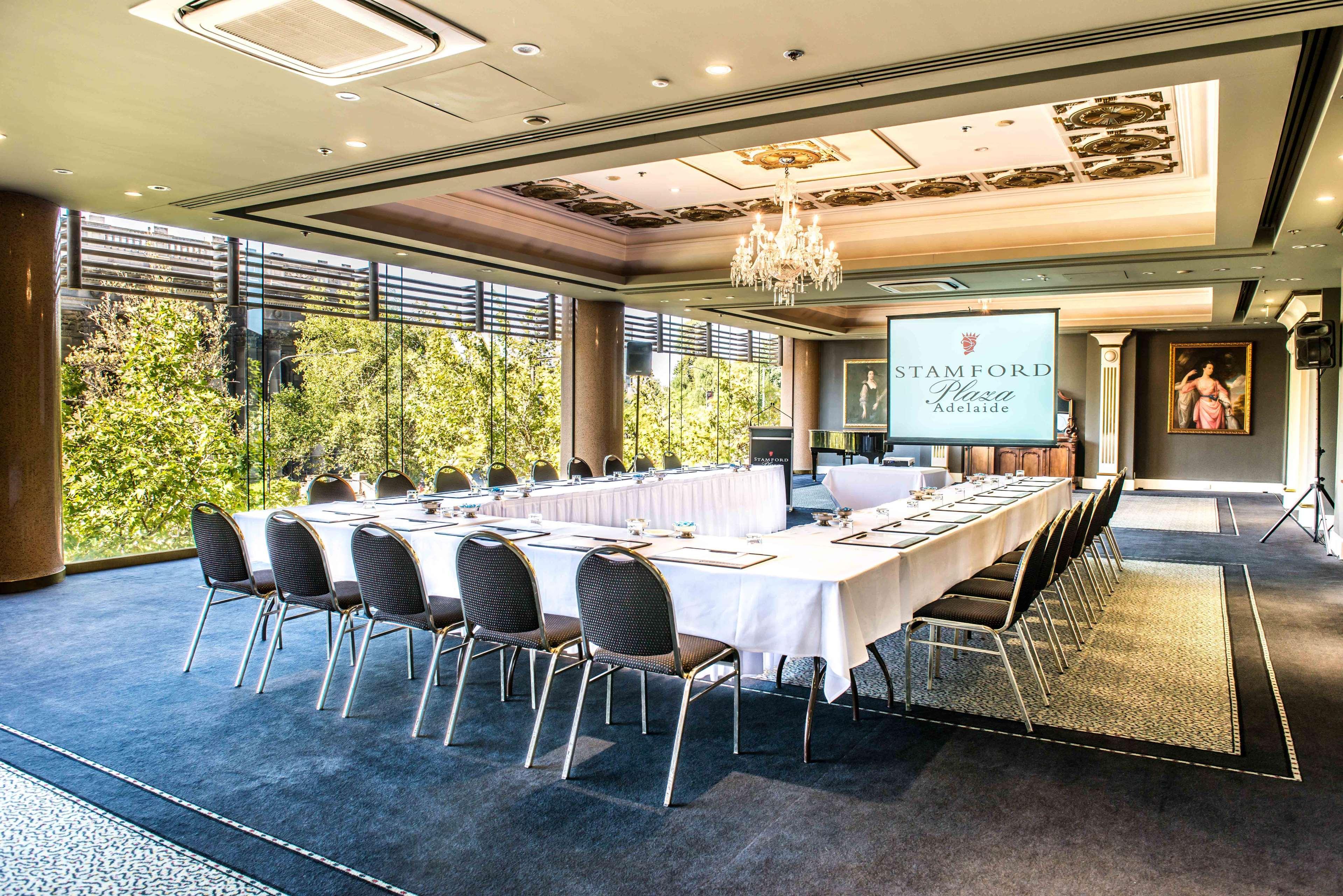 스탬퍼드 플라자 애들레이드 호텔 외부 사진 The photo shows a stylish conference room set up for a meeting or event. There is a long rectangular table covered with a white tablecloth, surrounded by black chairs. A large screen displays the name "Stamford" along with a logo. The room features l